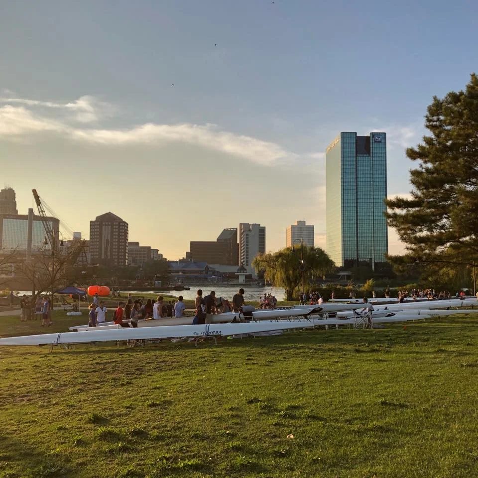 Shells and rowers gathered at the boathouse at sunset with the Toledo skyline behind