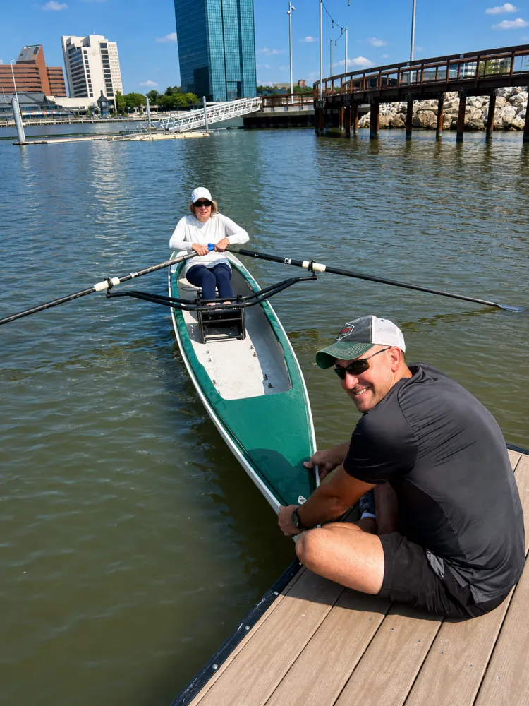 A coach steadying a single scull at the dock as a rower prepares to launch on the Maumee River
