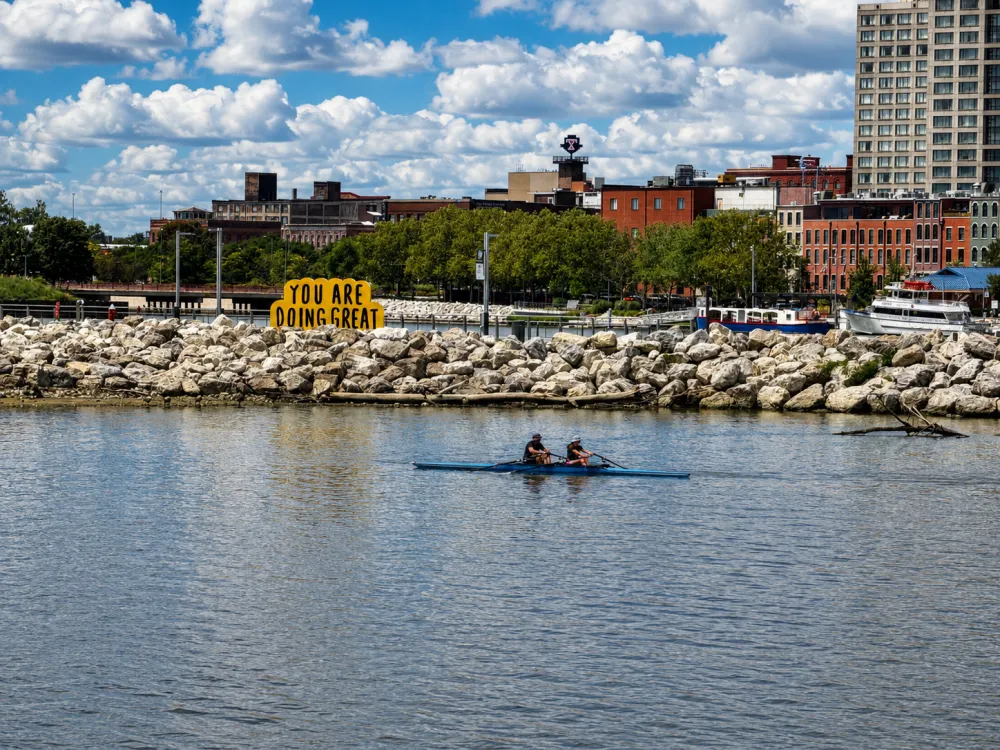 A double scull gliding past a "You Are Doing Great" sign on the Maumee River in downtown Toledo